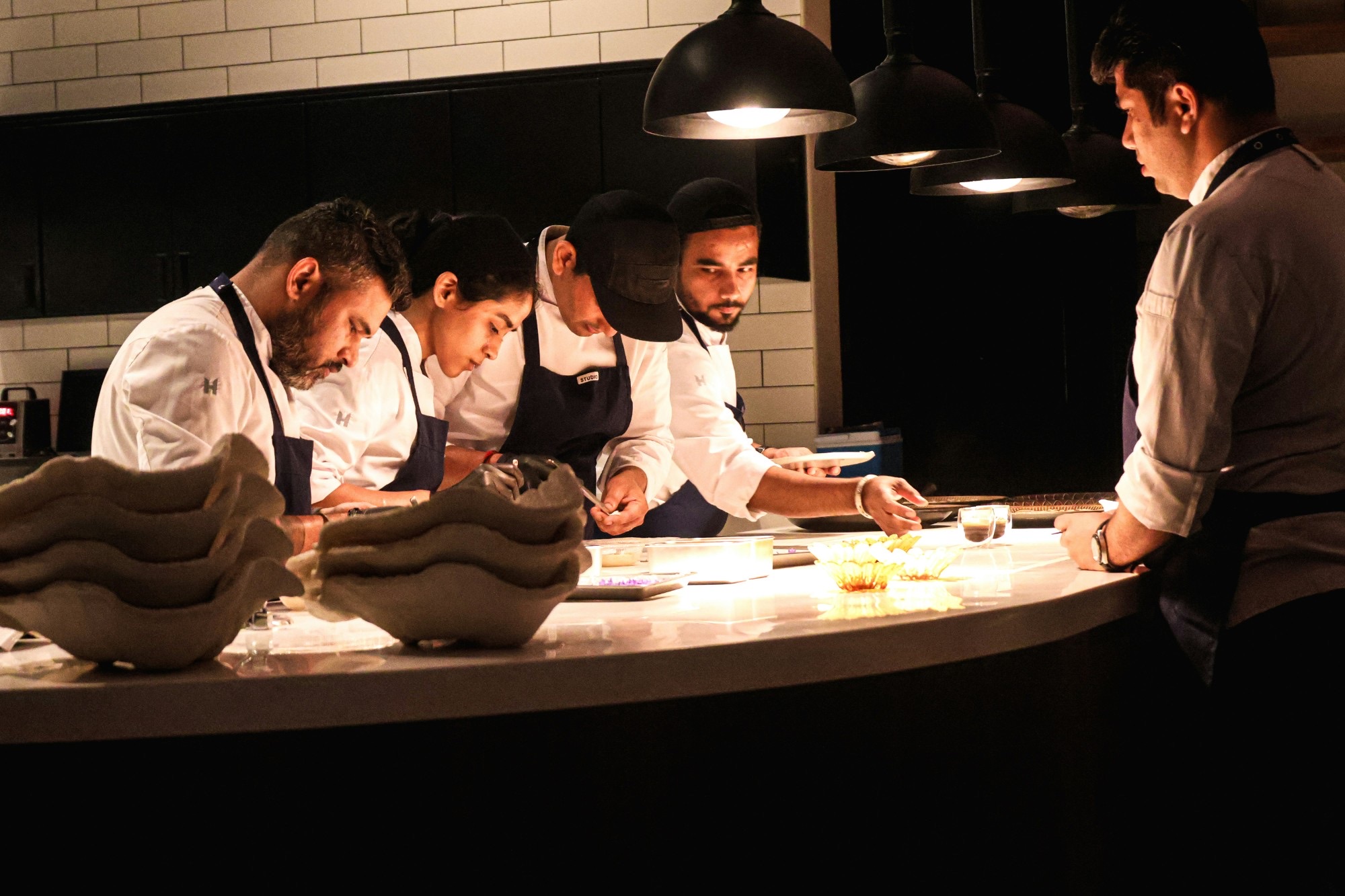 A kitchen team plating at the pass under pendant lamps while the head chef watches